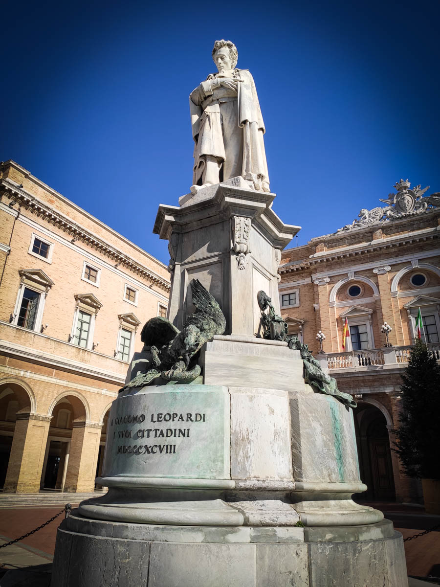 statua marmorea di Giacomo Leopardi su alto basamento con figure allegoriche in bronzo, Piazza Leopardi a Recanati