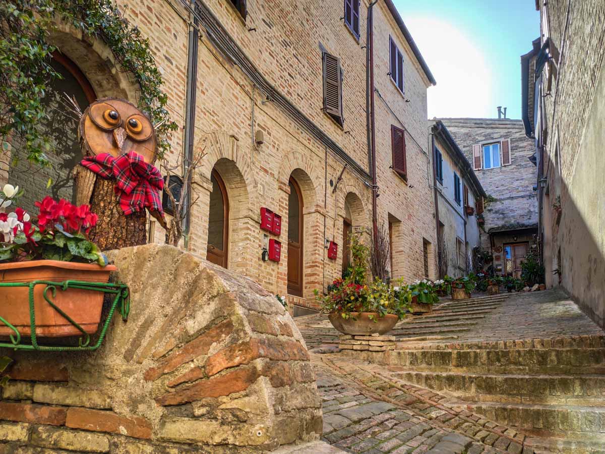 vicolo di Recanati con scalinata in mattoni e una civetta scolpita decorata con fiori rossi