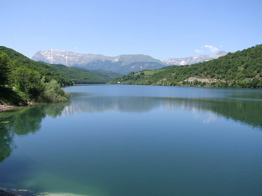 il Lago di Gerosa con l'acqua blu e le sponde boscose, sullo sfondo il Monte Vettore e il Monte Torrone ancora innevati