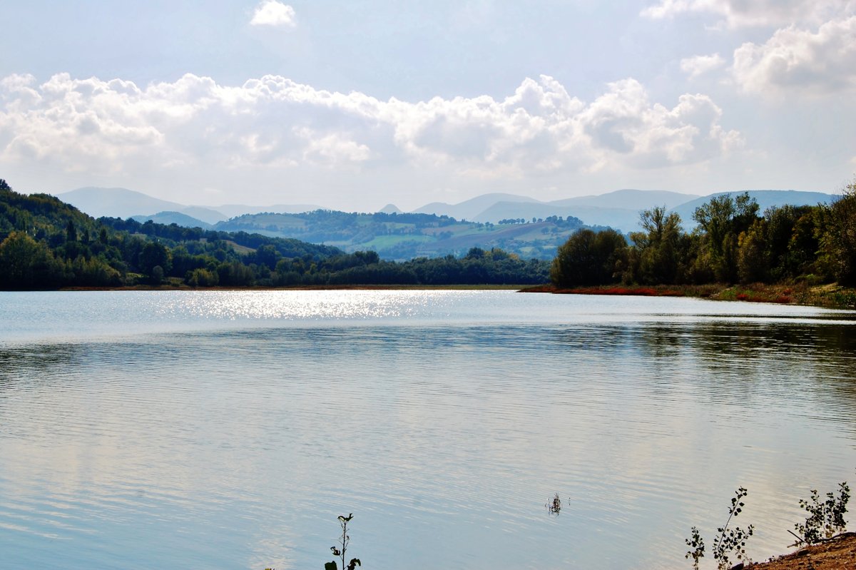 il Lago di Polverina con l'acqua calma che riflette il sole, sponde boscose e le colline dell'Appennino all'orizzonte
